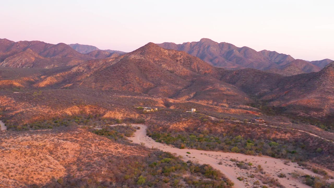 Aerial view in Cabo Pulmo, Baja California Sur with warm light on dry foreground terrain, a pale road bending through scrub, small structures near the midground, mountains behind, and no visible people.