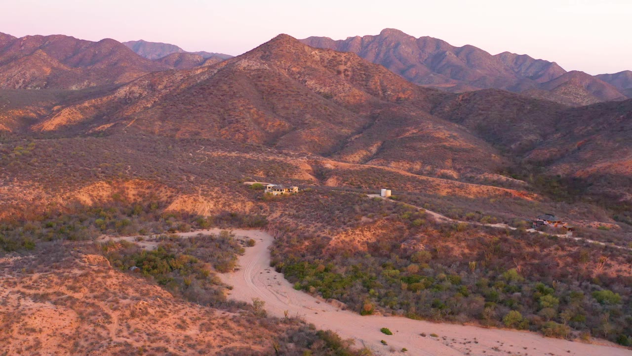 Cabo Pulmo desert valley seen from above, with the winding road lower in frame, scrub vegetation across the hills, small structures in the distance, and no visible people.