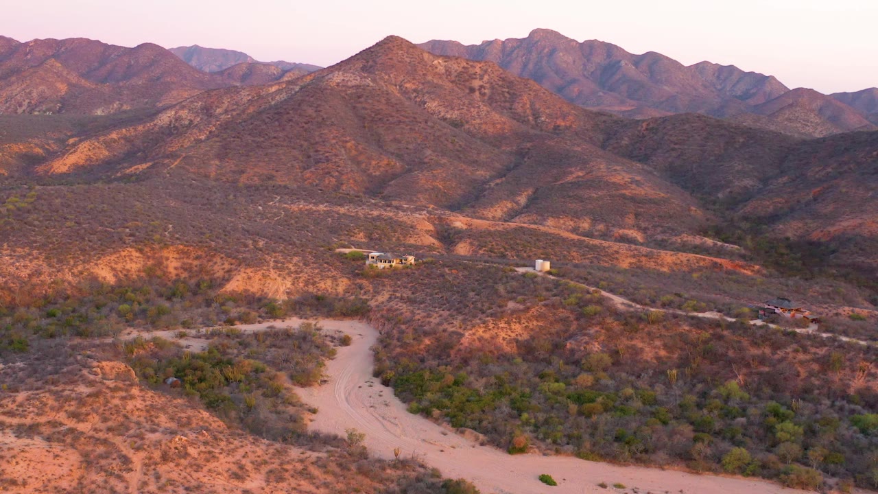 Aerial view across dry hills toward a mountain range and winding road.