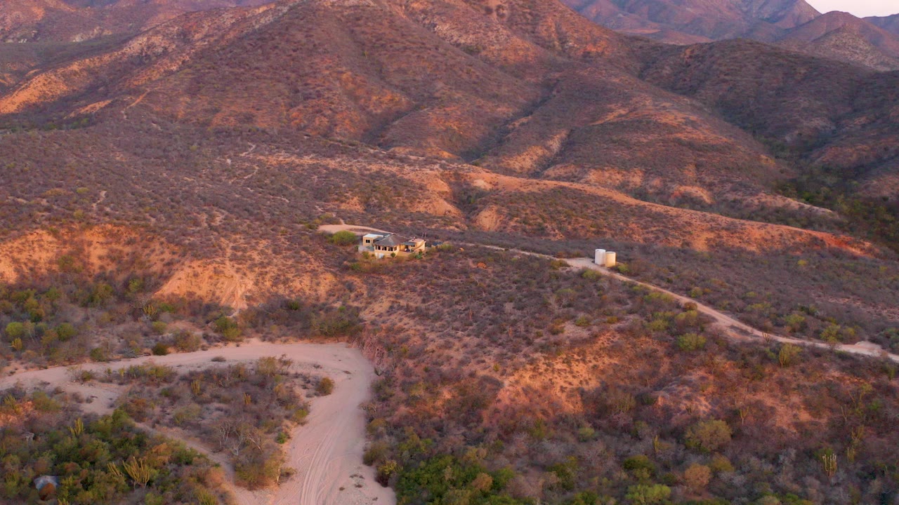 Aerial view over Cabo Pulmo desert hills with pale dirt roads, scattered shrubs, a small structure, white tanks, layered mountains, and no visible people.