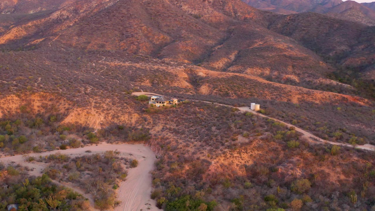 Aerial frame over Cabo Pulmo desert terrain with reddish sunlit slopes, patchy shrubs, a pale road, a small structure, white tanks, and no visible people.