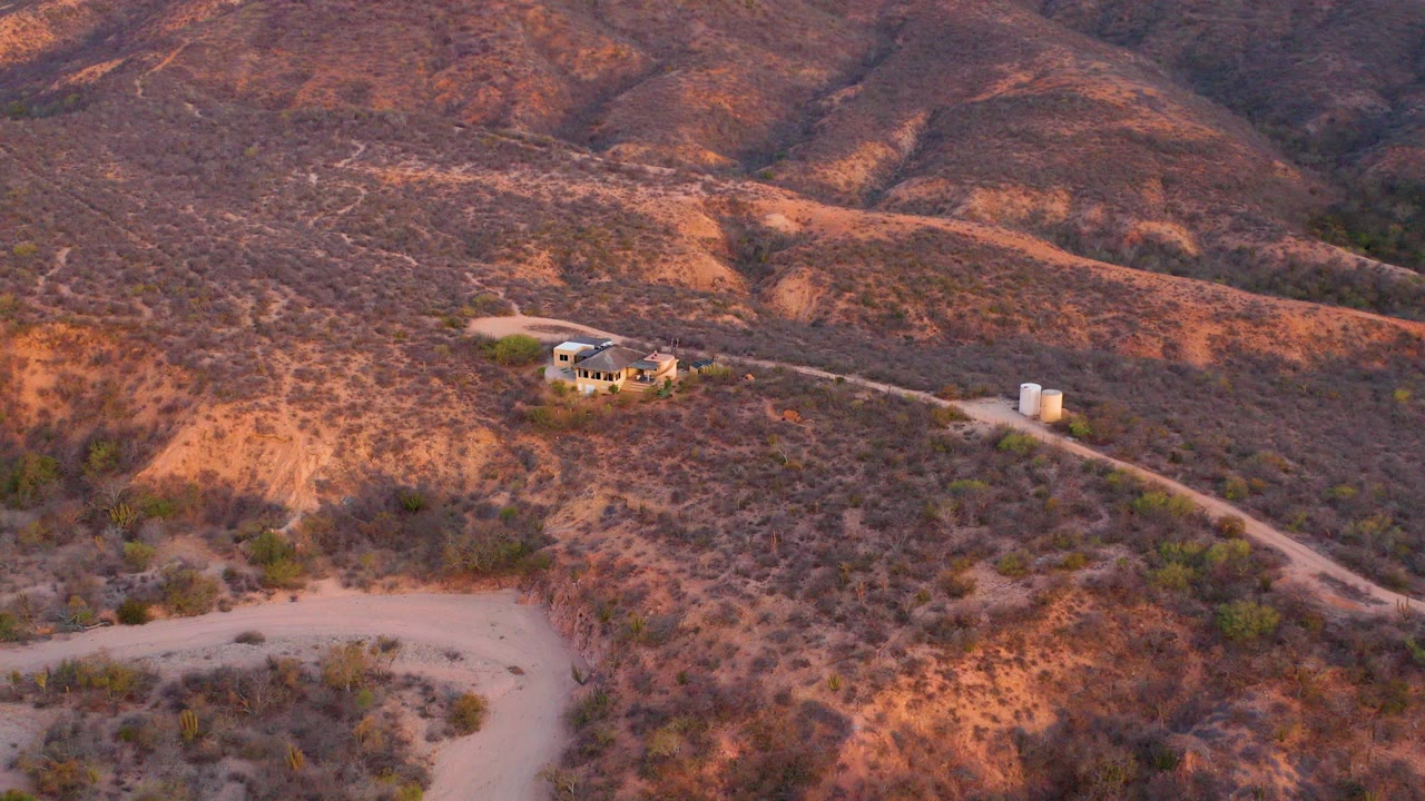 High aerial view in Cabo Pulmo, Baja California Sur showing a small hillside house, winding dirt road, white cylindrical tanks, dry brush, and no visible people.