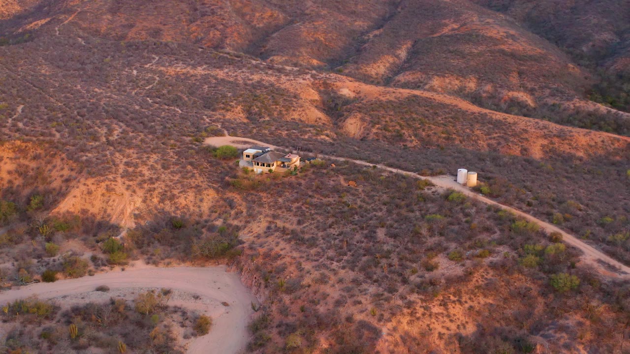 Cabo Pulmo aerial view showing the hillside house slightly left of center, the dirt access road crossing toward two cylindrical tanks, warm ridge light, and no visible people.