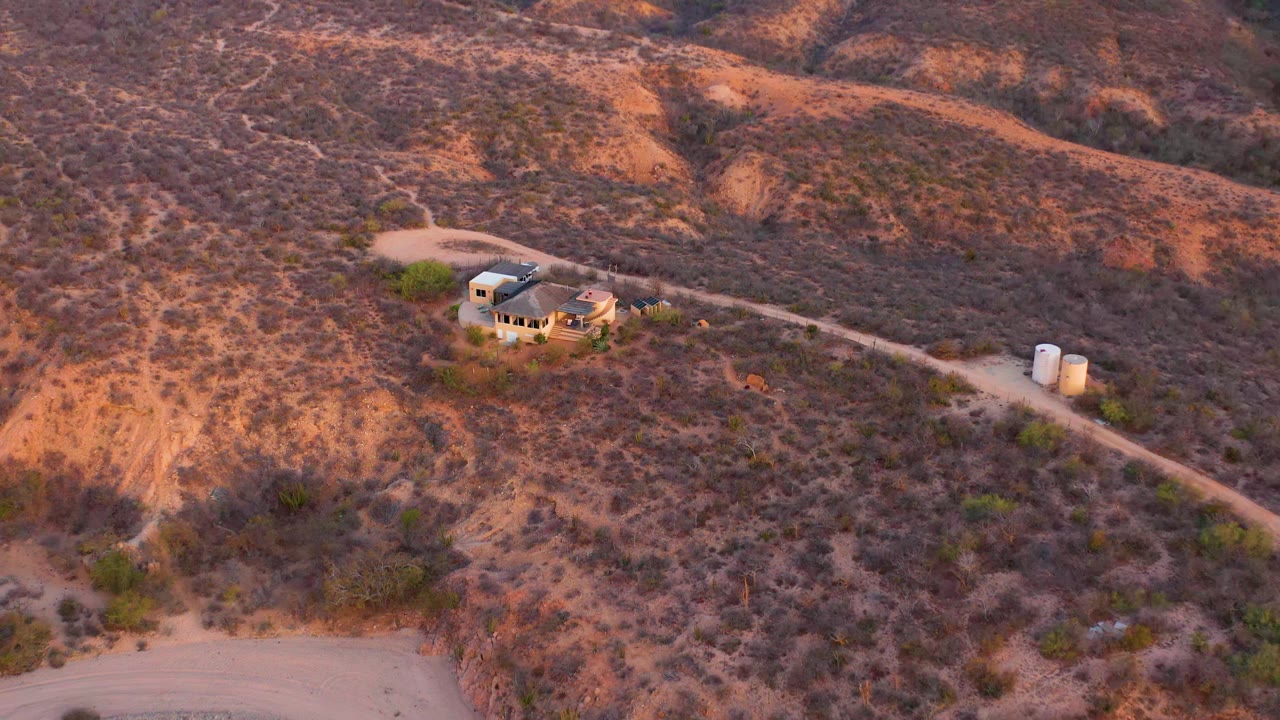 High oblique view in Cabo Pulmo, Baja California Sur with the house left of center, two tanks near the road, orange-brown hills, and no visible people.
