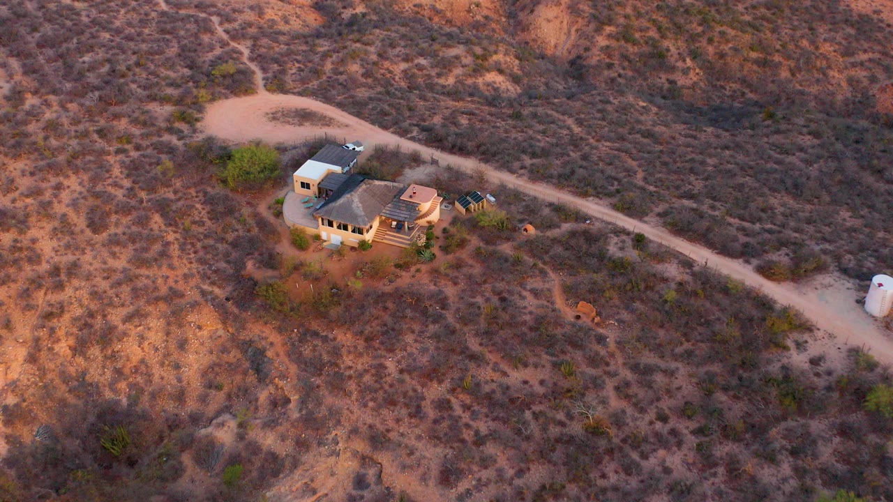 Oblique aerial frame over a house compound and dirt road in Cabo Pulmo, Baja California Sur, with dry scrubland around it and no visible people.