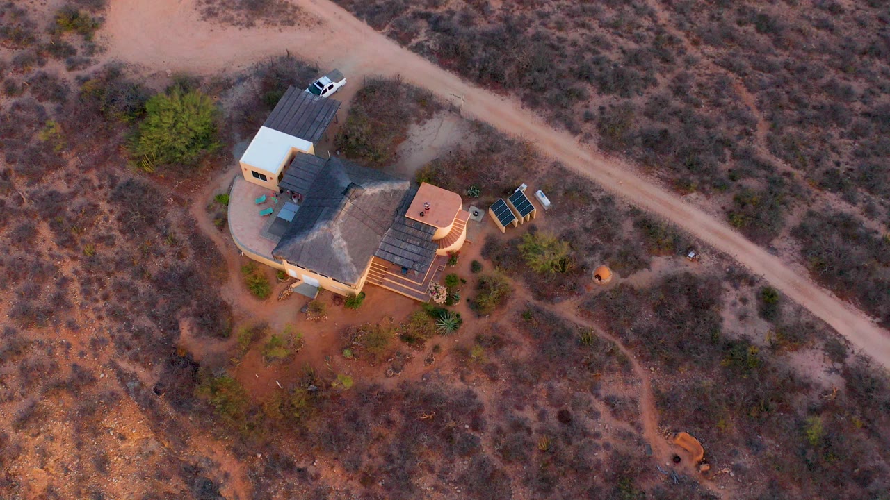 Overhead view in Cabo Pulmo, Baja California Sur, showing a rural house compound beside a pale dirt road, with dry brush around it and no visible people.