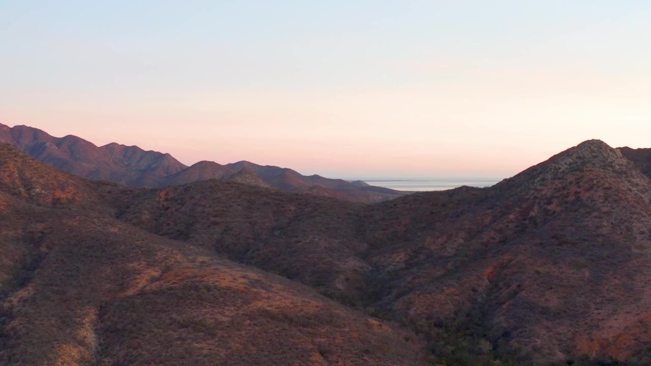 Rugged Cabo Pulmo hills run across the foreground, with darker mountains and a narrow strip of distant water visible beyond them, with no visible people.