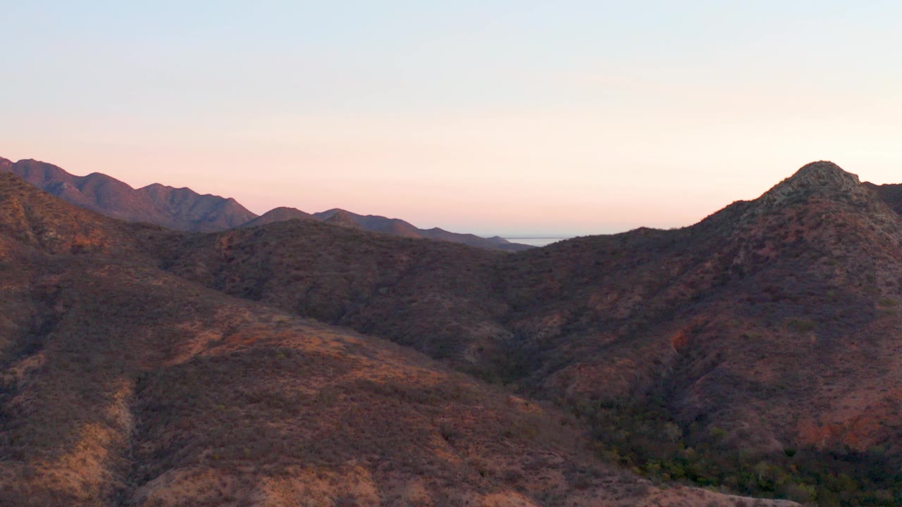 Wide aerial view of Cabo Pulmo mountain ridges at dusk with dry slopes, distant peaks, pale pink sky, and no visible people.