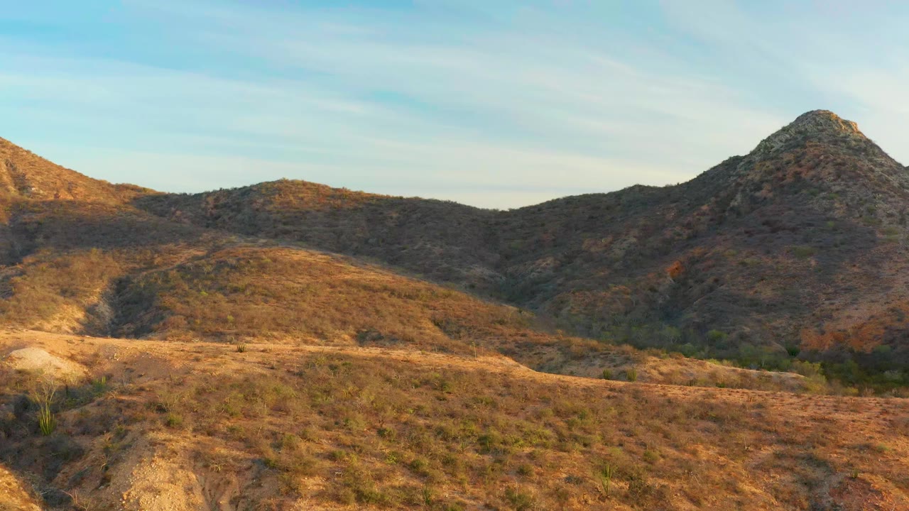 Cabo Pulmo hillside terrain with brown grasses, scrub, a rocky peak near the right side, and no visible people beneath a lightly clouded sky.