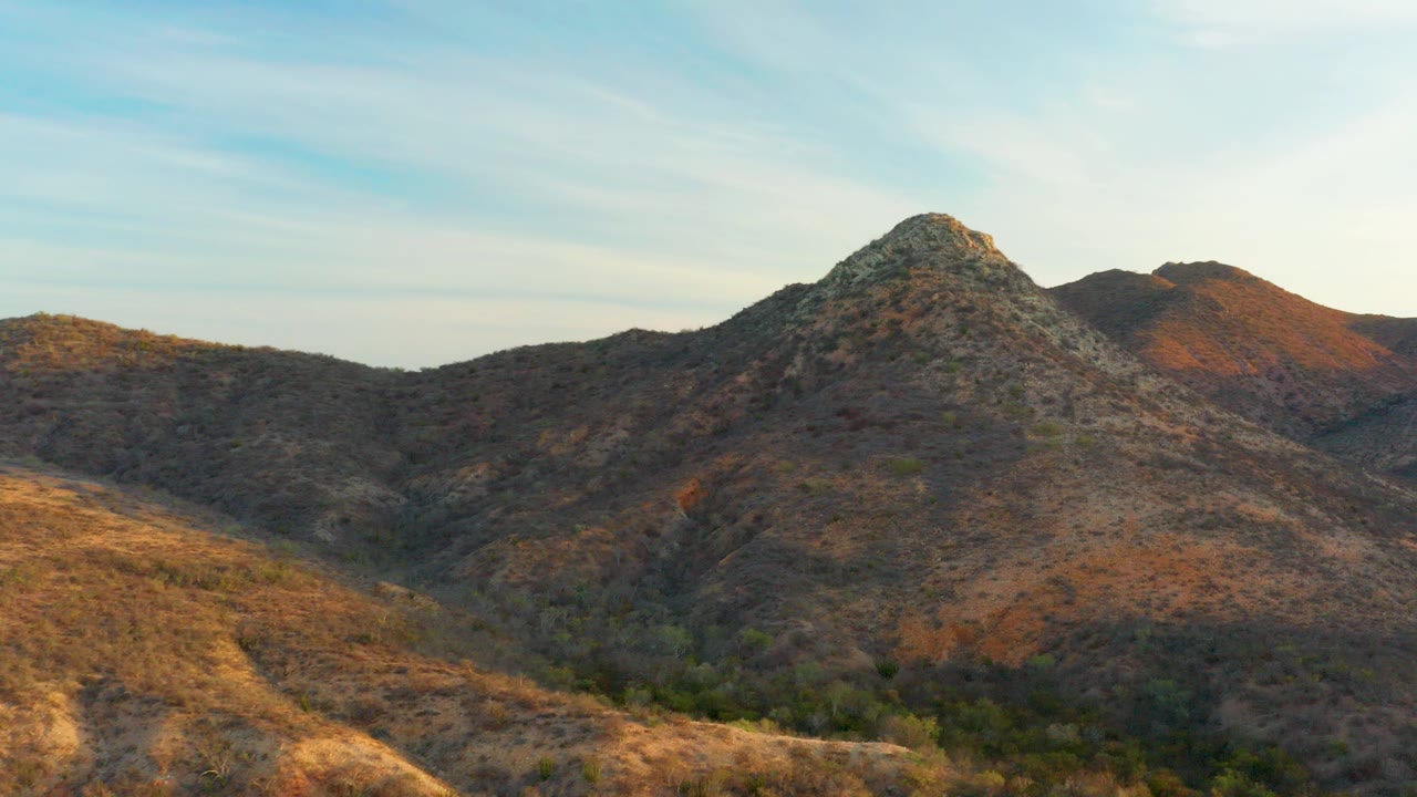 Cabo Pulmo, Baja California Sur aerial frame with a rocky central peak, dry brown slopes, green brush in the lower valley, thin clouds, and no visible people.