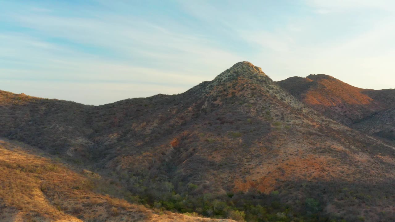 Cabo Pulmo aerial frame with dry ridges crossing the foreground, a craggy peak near center, orange-brown light on the right hillside, and no visible people.