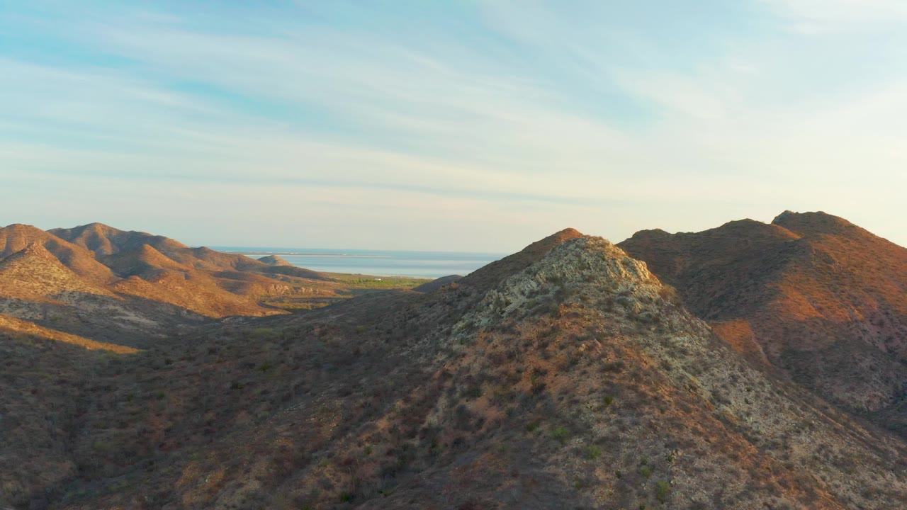The aerial composition looks across dry Cabo Pulmo hills toward a distant blue waterline, with the rocky foreground ridge lower in frame and no visible people.