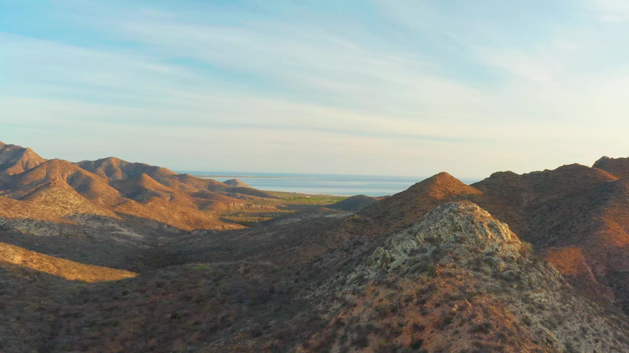 Sunlit arid ridges in Cabo Pulmo stretch across the frame, with a shadowed valley below, distant ocean, thin clouds, and no visible people.