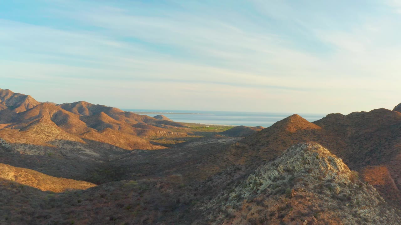 A dry Cabo Pulmo valley sits between low rocky mountains, with warm light on the ridges, distant water at the horizon, and no visible people.