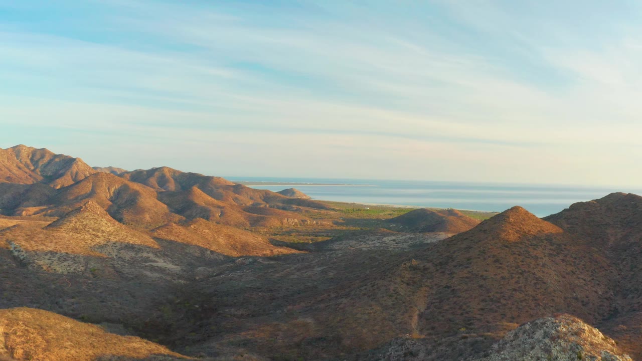 A high view across Cabo Pulmo desert hills shows sunlit slopes, a darker foreground ridge at right, distant water, and no visible people.