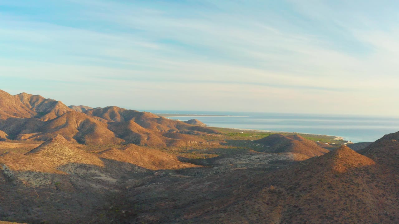 Wide view of arid Cabo Pulmo mountain ridges with the ocean and low coastline in the distance under a pale streaked sky, no visible people.