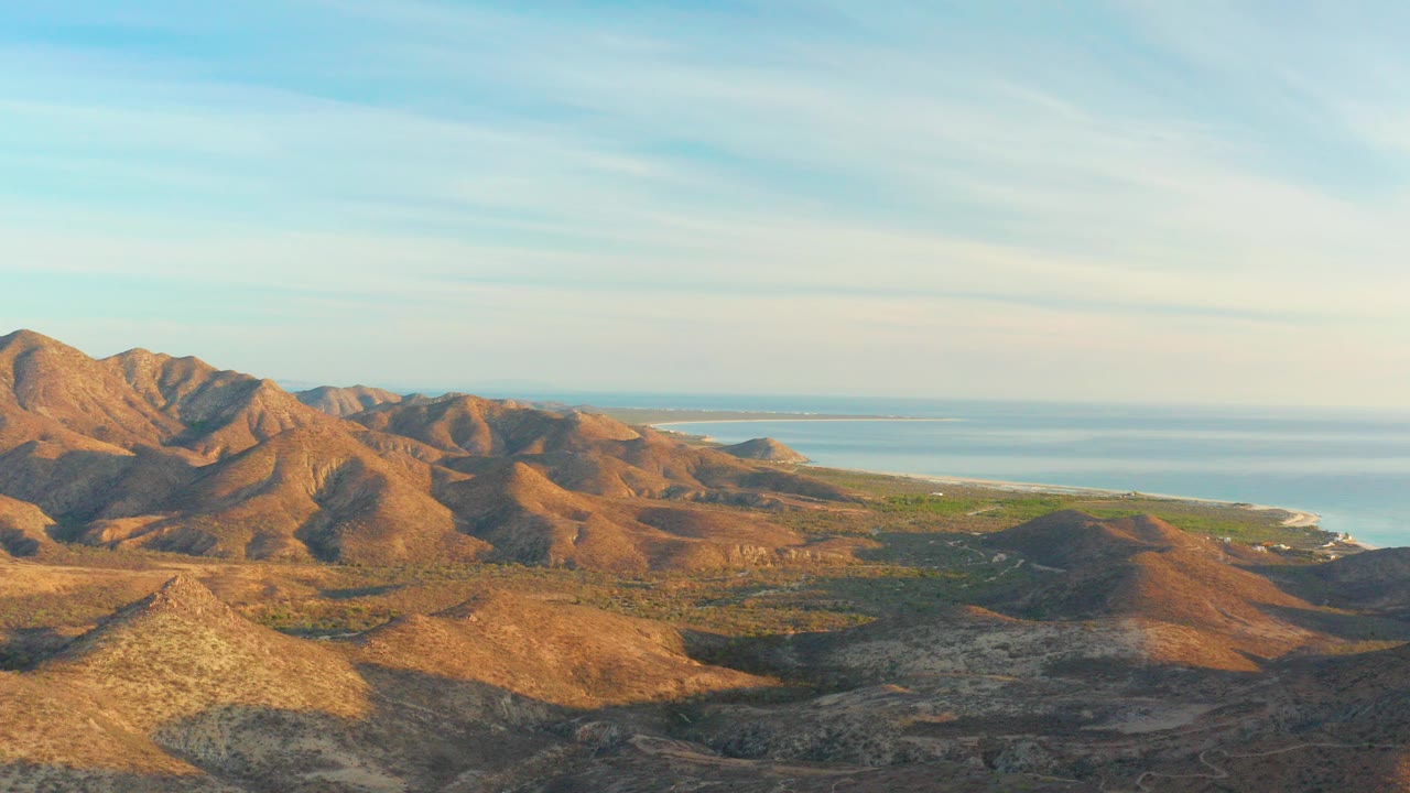 Wide aerial view of Cabo Pulmo mountain ridges with sparse green valley areas, pale blue ocean on the horizon, and no visible people.
