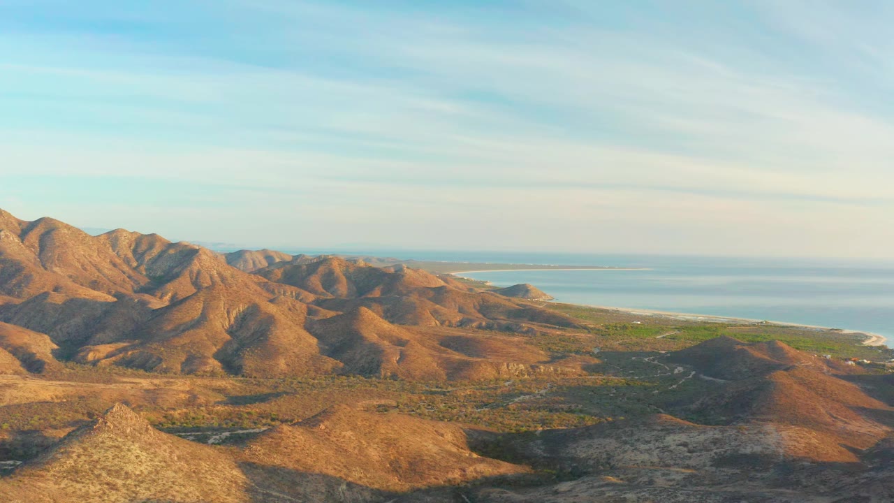 Cabo Pulmo hills fill the foreground and midground, with warm sunlight on brown slopes, darker valley shadows, a narrow coastal strip, distant ocean, and no visible people.