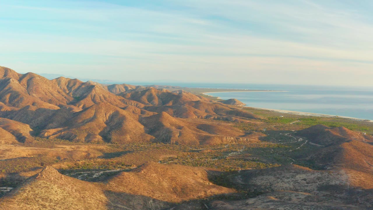 Opening frame of Cabo Pulmo, Baja California Sur mountains under warm light, with dry ridges in front, blue sea beyond, thin clouds overhead, and no visible people.