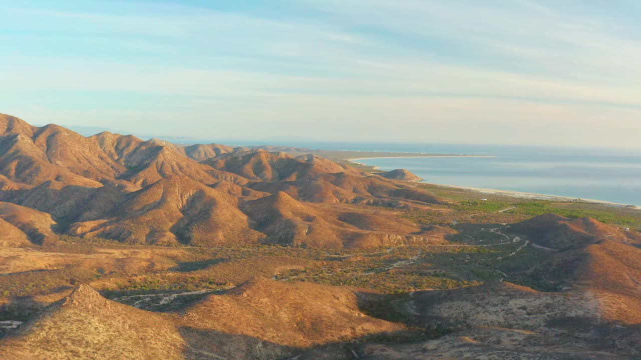 Cabo Pulmo mountain ridges remain spread across the frame, with warm sunlight on tan slopes, darker valleys below, the sea at upper right, and no visible people.