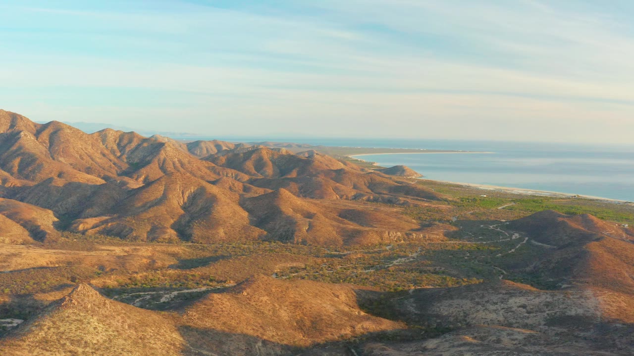 A broad aerial frame shows the Cabo Pulmo mountains slightly deeper in warm light, with layered hills, a blue waterline behind them, soft cloud bands, and no visible people.