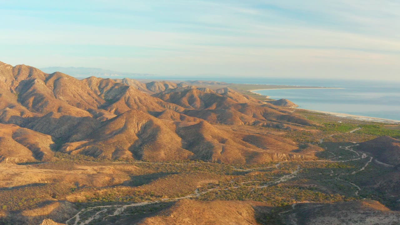 Aerial frame over Cabo Pulmo showing golden dry hills, a winding road through low vegetation, blue ocean beyond the mountains, and no visible people.