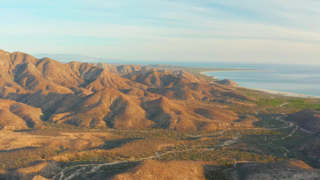 Aerial view in Cabo Pulmo with sunlit brown ridges across the left side, pale road tracks crossing the valley floor, ocean in the distance, and no visible people.
