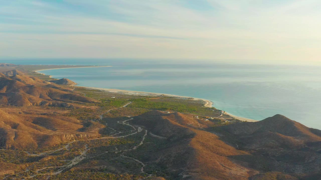 Early frame of Cabo Pulmo, Baja California Sur, with brown hills in the foreground, pale roads leading toward a sandy beach, blue ocean beyond, and no visible people.