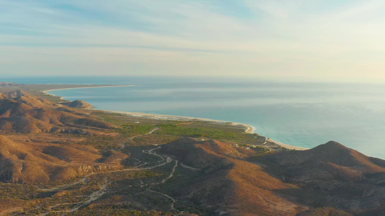 Cabo Pulmo aerial frame with the road more centered through dry brown terrain, green vegetation near the coast, pale shoreline, blue sea, and no visible people.