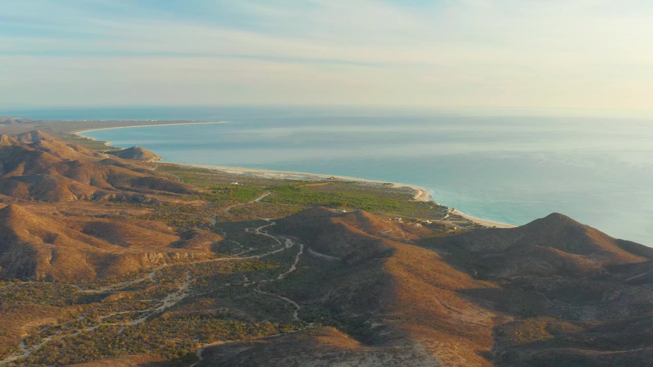 High aerial frame over Cabo Pulmo showing sunlit brown ridges, green lowland vegetation, a curving shoreline, calm blue water, and no visible people.