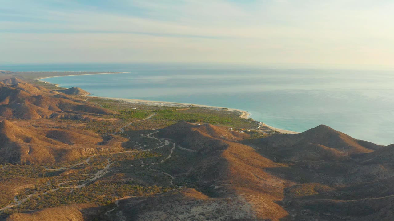 Elevated Cabo Pulmo view with warm light on brown hills, winding tracks through the lower terrain, a green shoreline band, and open blue water, with no visible people.