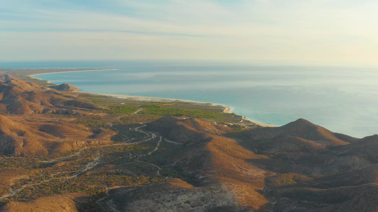 Mid-clip view of Cabo Pulmo with a dark hill in the lower right, ocher slopes across the middle, a green coastal plain, and pale surf line against blue ocean, with no visible people.