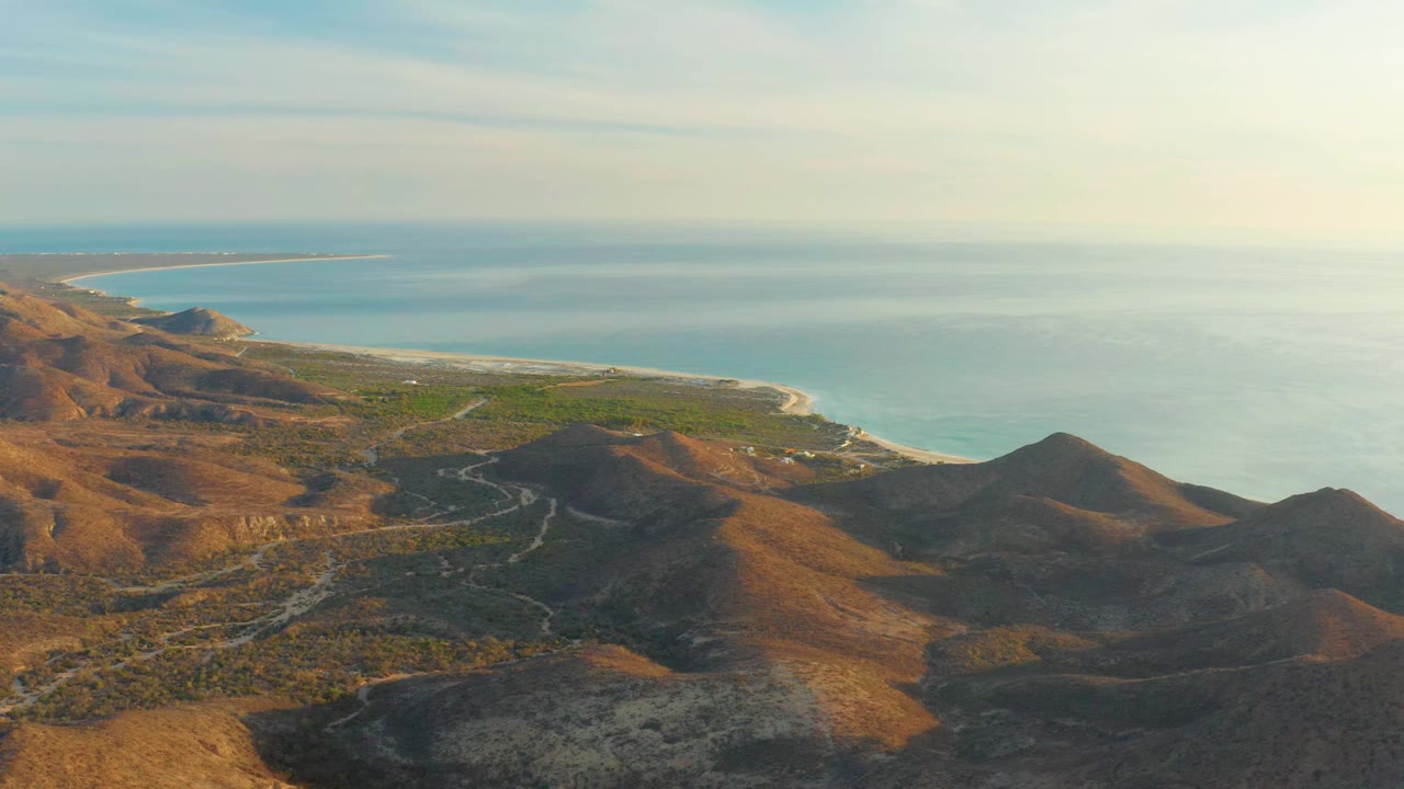 Aerial frame of Cabo Pulmo desert hills with scattered green vegetation, winding tracks, a light beach edge, blue ocean, and no visible people.
