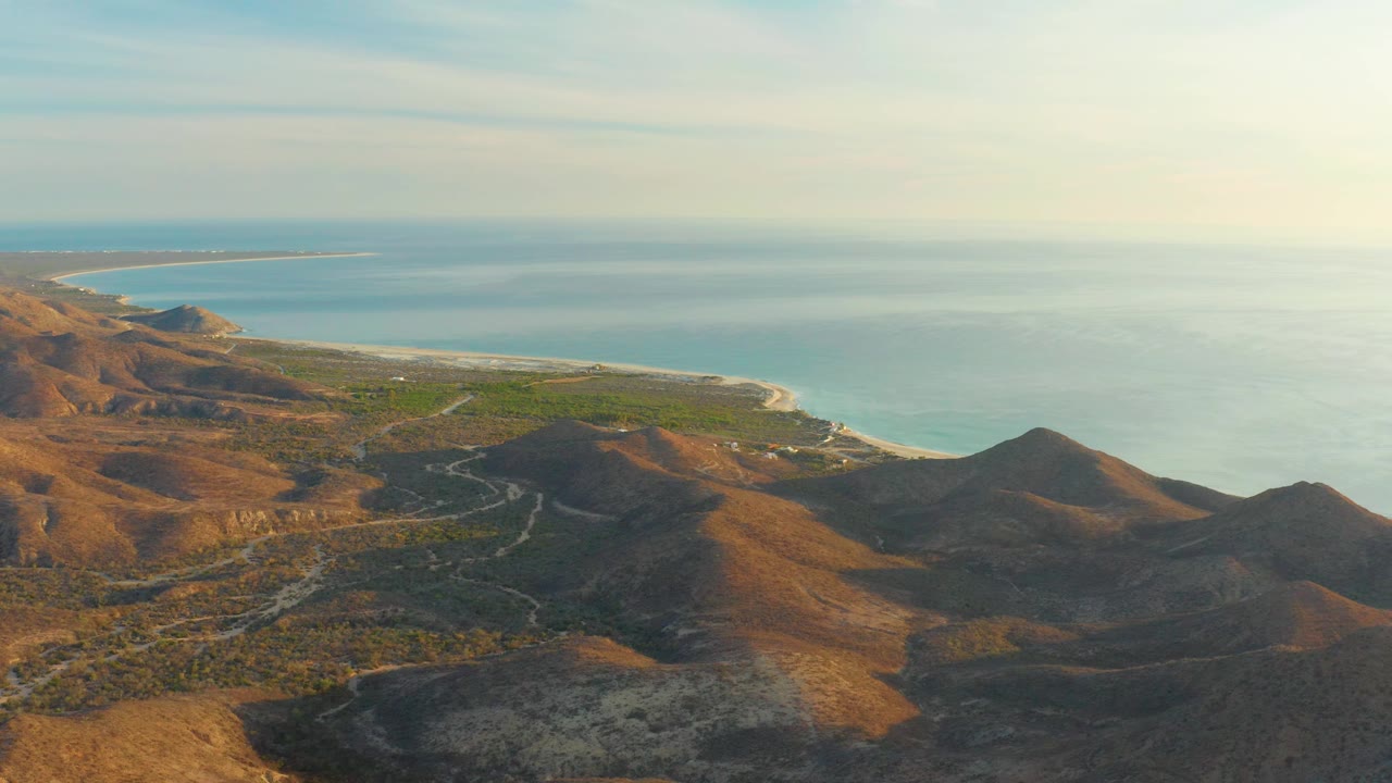 High aerial view in Cabo Pulmo showing sunlit brown slopes, darker ridge shadows, a narrow beach line, calm blue ocean, and no visible people.