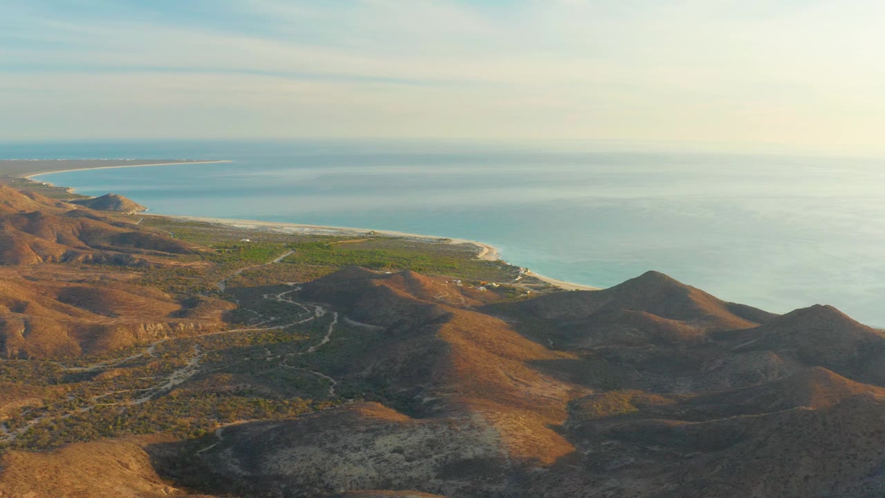 Cabo Pulmo aerial landscape with foreground hills in partial shadow, a green lowland near the beach, ocean in the background, and no visible people.