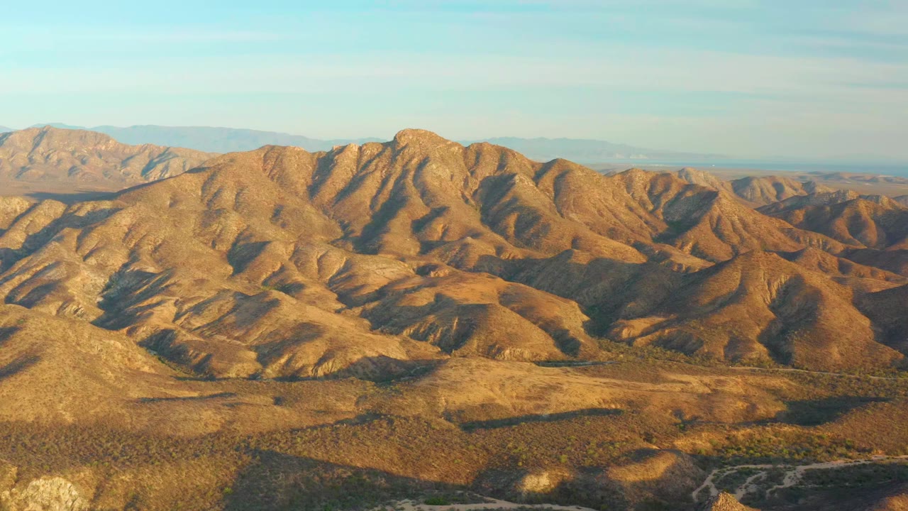 Aerial frame of Cabo Pulmo arid ridges with golden light across the slopes, darker valley folds, a pale horizon, and no visible people.