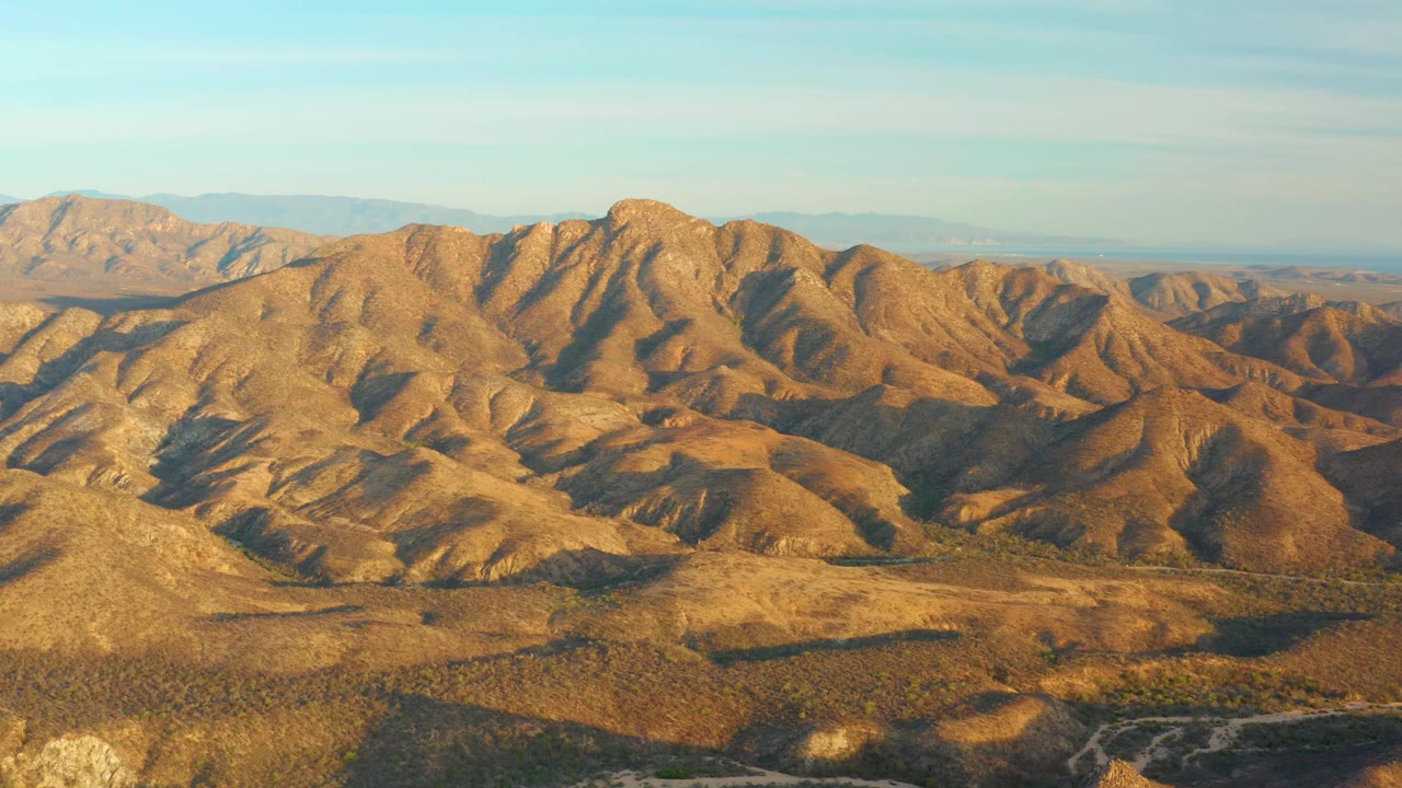 Cabo Pulmo mountain ridges fill the frame, with long warm shadows crossing dry valleys and a thin blue sky above, with no visible people.