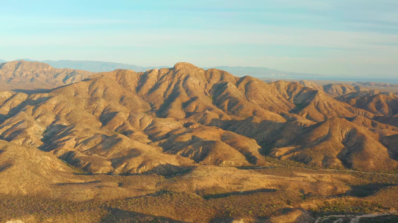 Golden arid ridges fill the frame near Cabo Pulmo, Baja California Sur, Mexico, with pale mountains on the horizon, a clear sky, and no visible people.