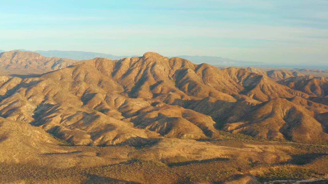 Rolling desert terrain near Cabo Pulmo, Baja California Sur, Mexico, shows sunlit slopes, shaded folds, a low band of distant mountains, clear sky, and no visible people.