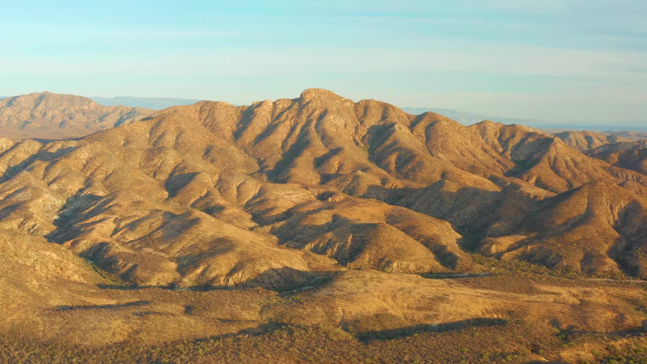 A high aerial frame over Cabo Pulmo desert hills, showing golden slopes, darker ravine shadows, sparse scrub, and no visible people.