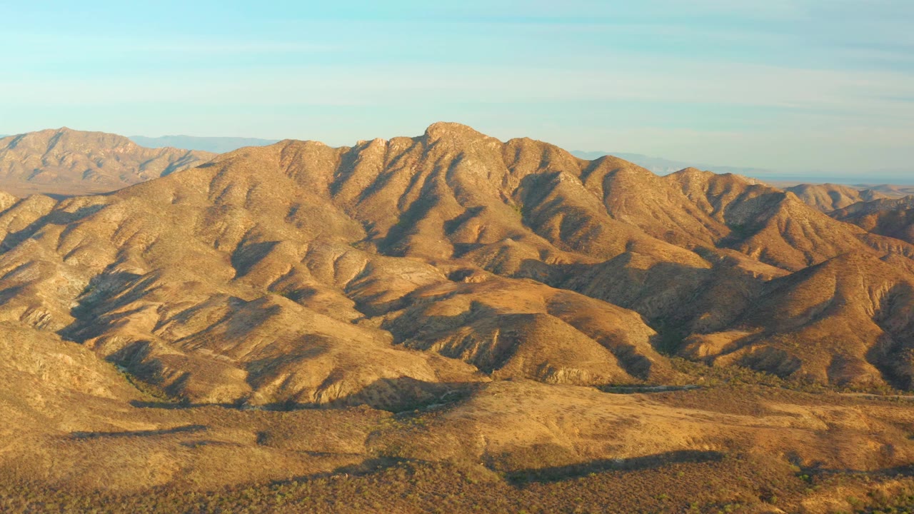 An elevated view across Baja California Sur desert hills shows warm brown slopes, deeper shadows in the folds, open sky, and no visible people.