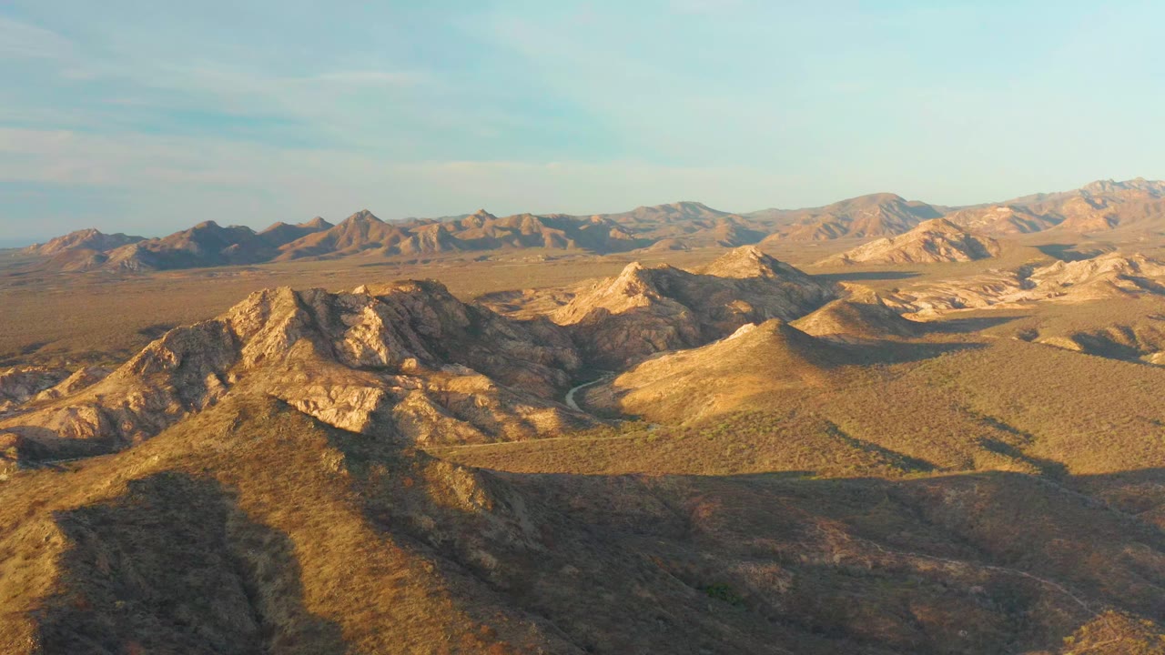Cabo Pulmo aerial frame with rocky foreground hills, a road bending through dry terrain, layered mountain ranges in the background, and no visible people.