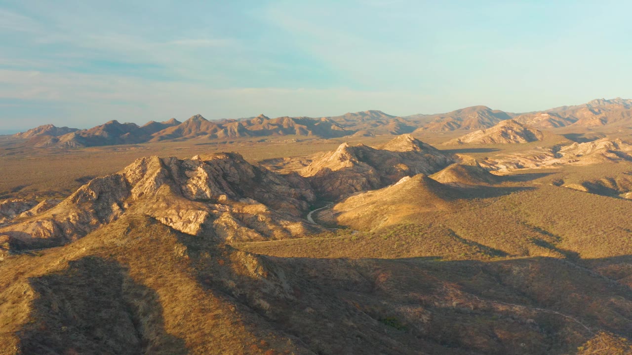 Cabo Pulmo aerial still showing a shadowed foreground slope, sunlit rocky ridges, the road curving through the basin, distant mountains, and no visible people.