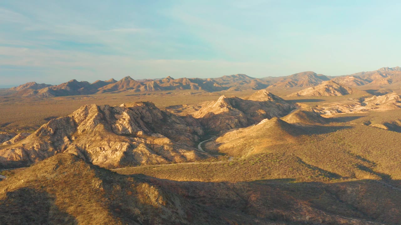 Aerial view over rocky desert hills near Cabo Pulmo, Baja California Sur, with a winding road in the valley, distant mountains, warm sunlight, and no visible people.