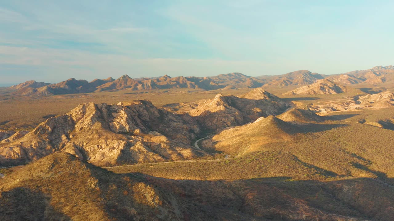 Aerial frame of Cabo Pulmo rocky hills in golden light, with shadowed slopes, a winding road crossing the open valley, distant mountains, and no visible people.