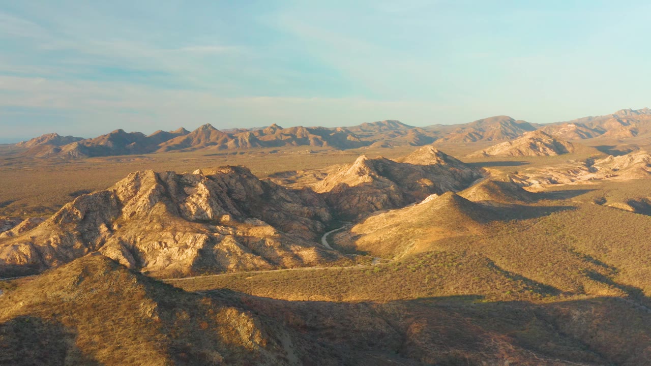 Wide aerial composition over Cabo Pulmo desert hills, showing a road threading between rocky slopes, broad dry terrain, a high blue sky, and no visible people.