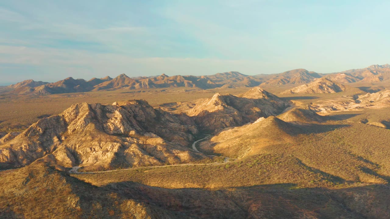 At one second, the Cabo Pulmo aerial view holds on sunlit tan ridges, a curving road in the valley floor, darker shadowed slopes, distant dry mountains, clear sky, and no visible people.
