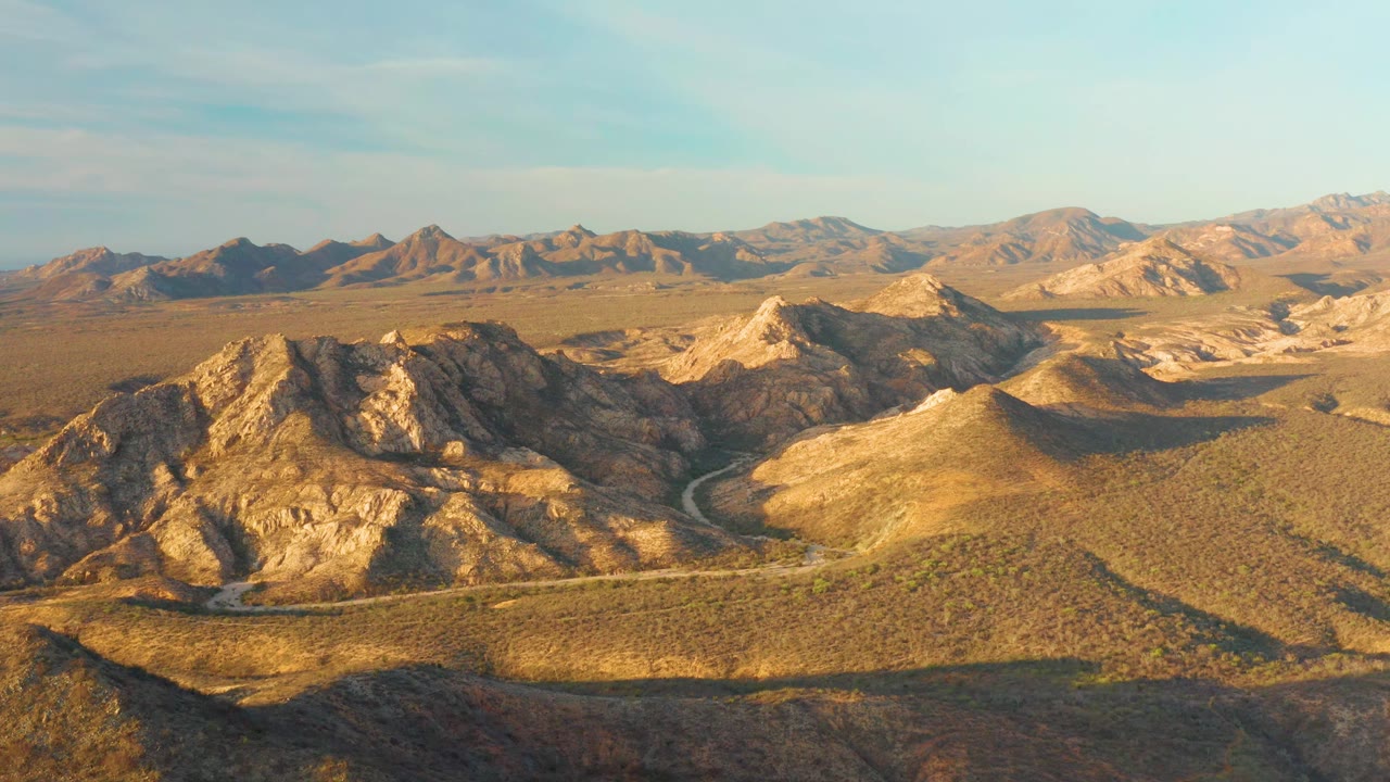Cabo Pulmo aerial frame with the road bend lower in the scene, a rugged ridge catching warm sun, distant brown mountains, clear blue sky, and no visible people.