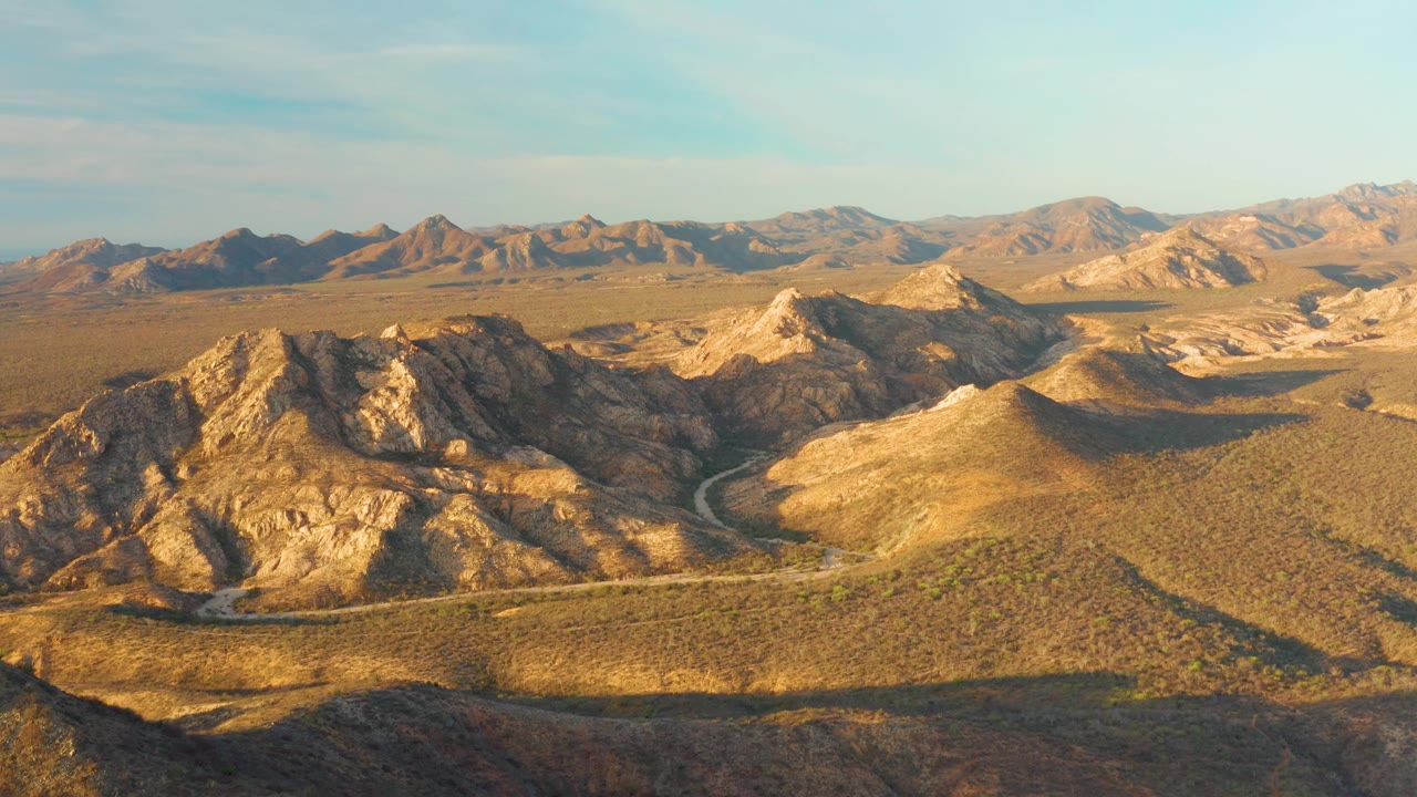 Cabo Pulmo drone-like frame with long shadows stretching across tan desert slopes, the pale road curving near the lower hills, distant mountains, and no visible people.
