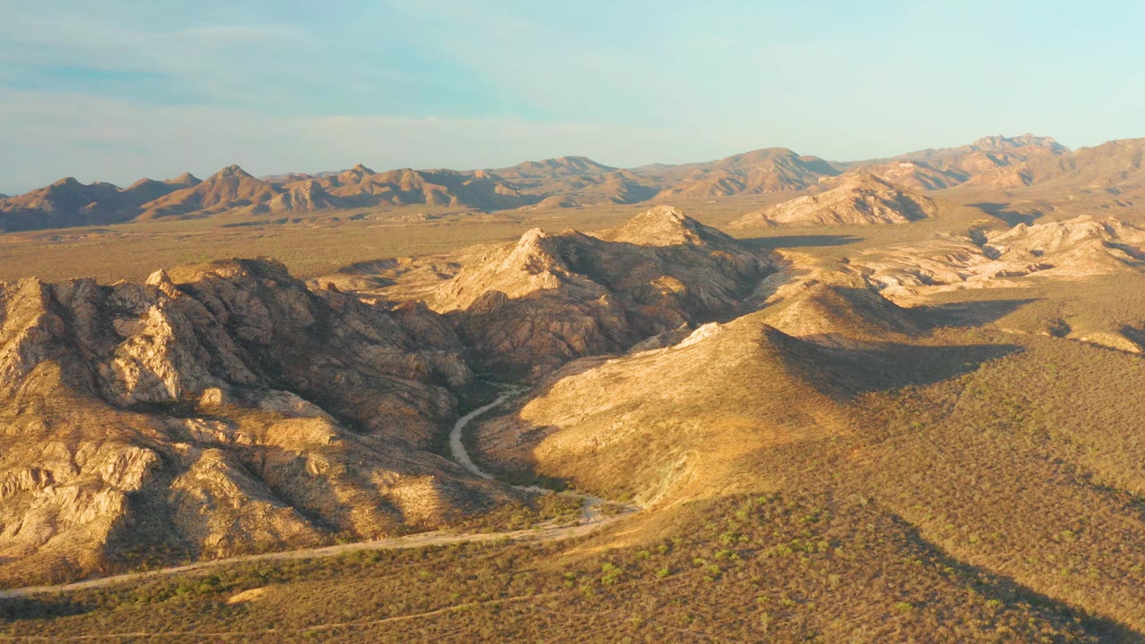 Aerial frame in Cabo Pulmo showing the winding dirt-colored road between sunlit desert slopes, darker rocky ridges, distant mountain ranges, and no visible people.